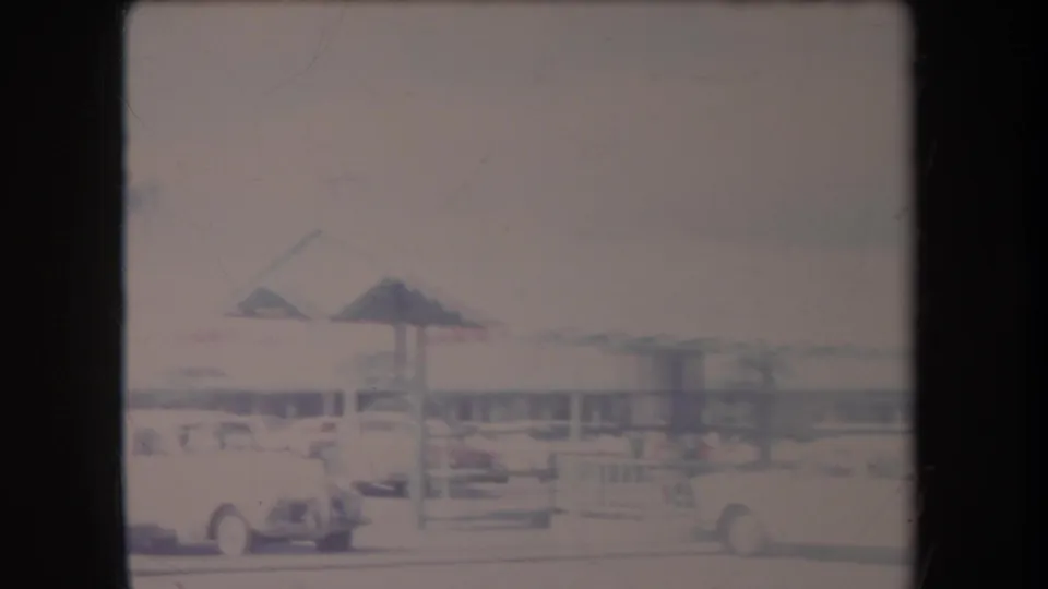 1969 Hawaii 8mm frame showing a low roadside building, parked cars, railings, and overexposed pavement in a commercial lot.