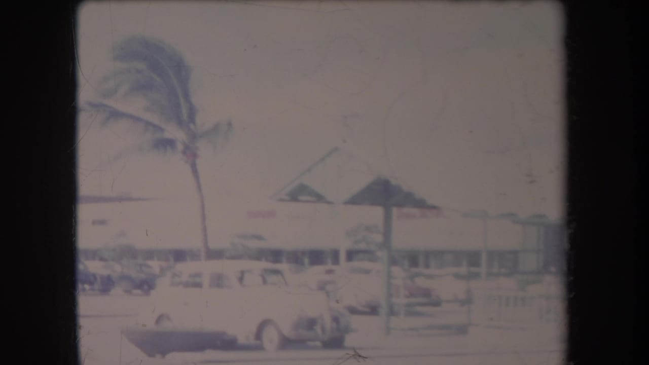1969 Hawaii home-movie frame showing palm trees, mid-century cars, and low commercial buildings along a bright street.