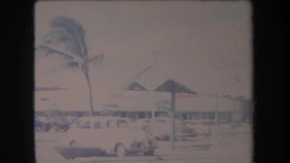1969 Hawaii home-movie frame showing palm trees, mid-century cars, and low commercial buildings along a bright street.