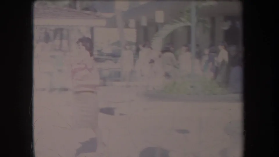 Soft 8mm frame of pedestrians near a Hawaii commercial sidewalk, with storefronts, posts, and street activity visible.