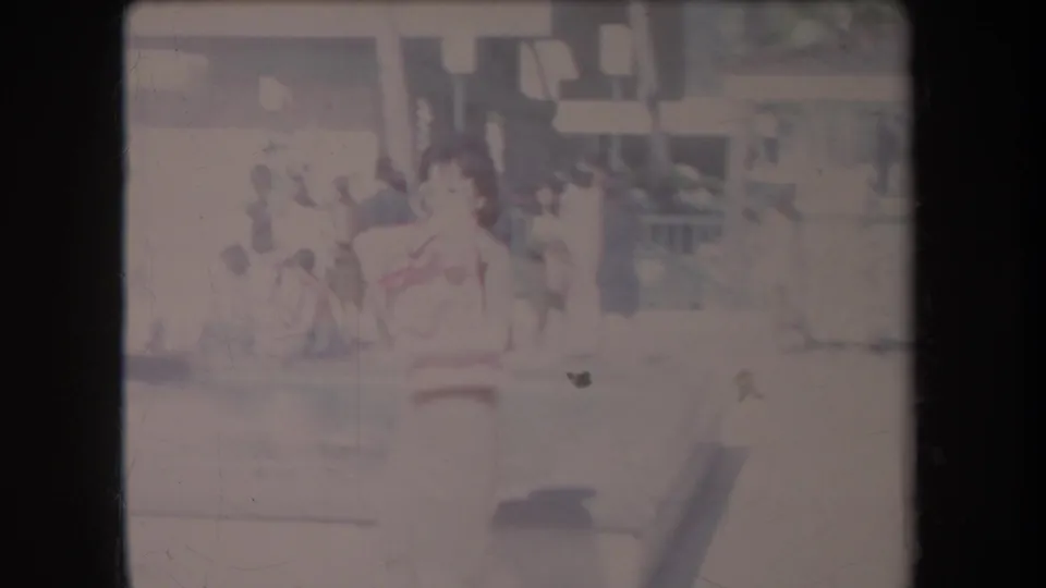 1969 Hawaii home-movie frame with a woman in light clothing near parked cars and pedestrians outside commercial buildings.