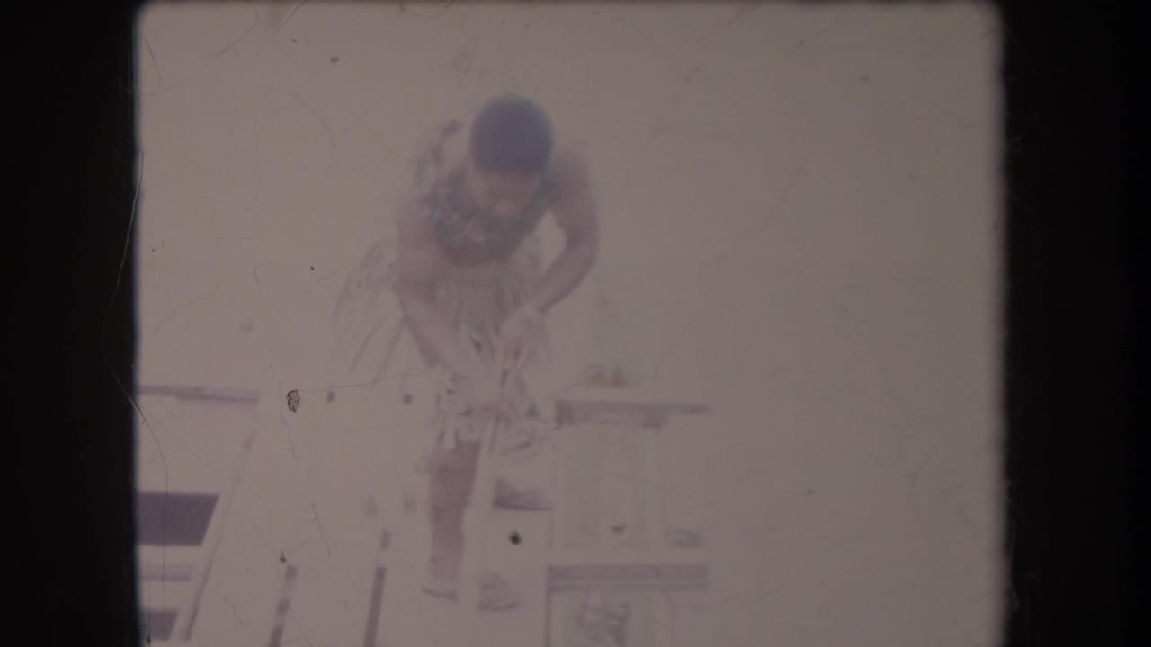 Shirtless man in a grass skirt bends on a wooden platform with a long pole nearby.