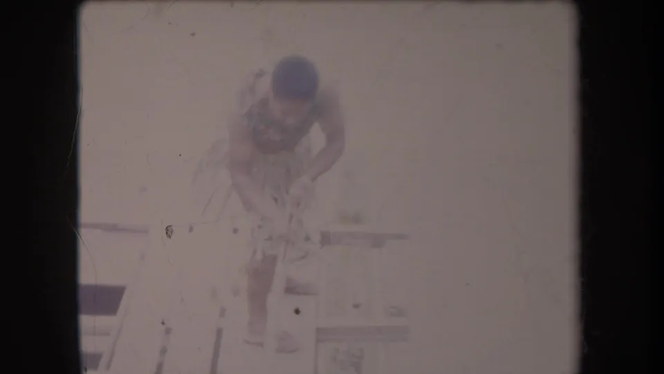 In faded 1969 Hawaii 8mm footage, a shirtless barefoot man in a grass skirt bends on a small wooden platform beside pale water.