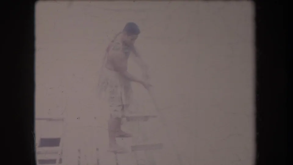 The man stands in profile on the wooden platform in Hawaii, holding a long pole for balance against an overexposed riverbank background.