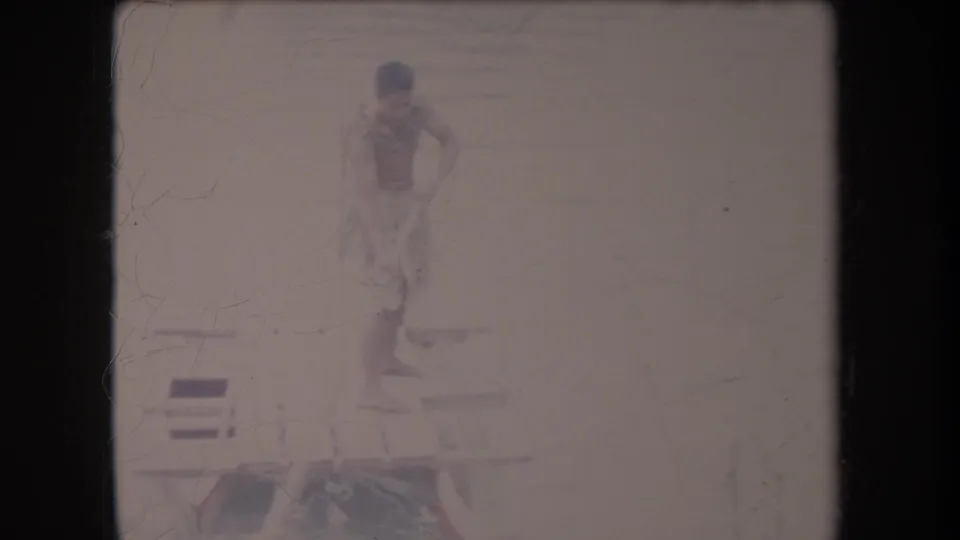 A wider faded frame shows the barefoot man in a grass skirt standing on the wooden platform, with water or riverbank visible behind him.