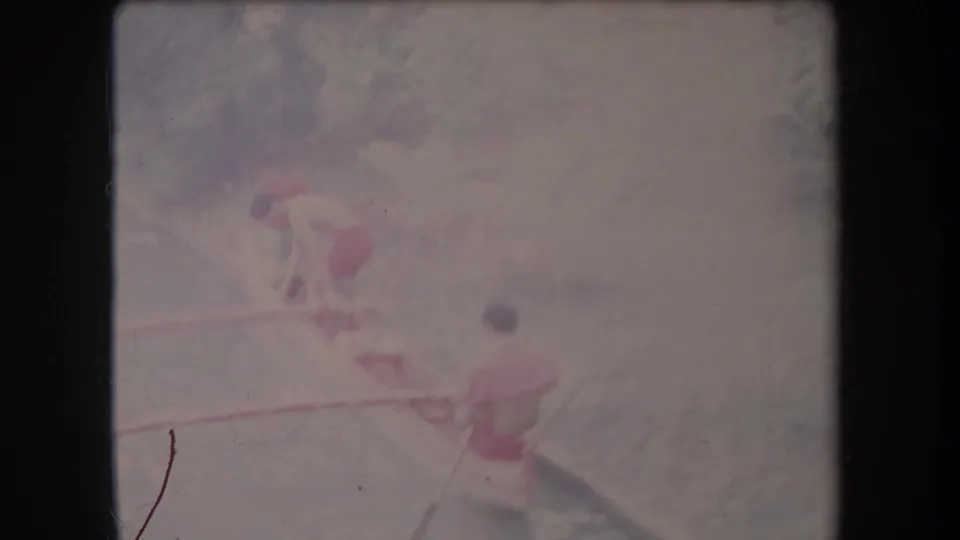 1969 Hawaii 8mm frame showing two visible men in casual swimwear paddling a narrow outrigger canoe on a tropical river.