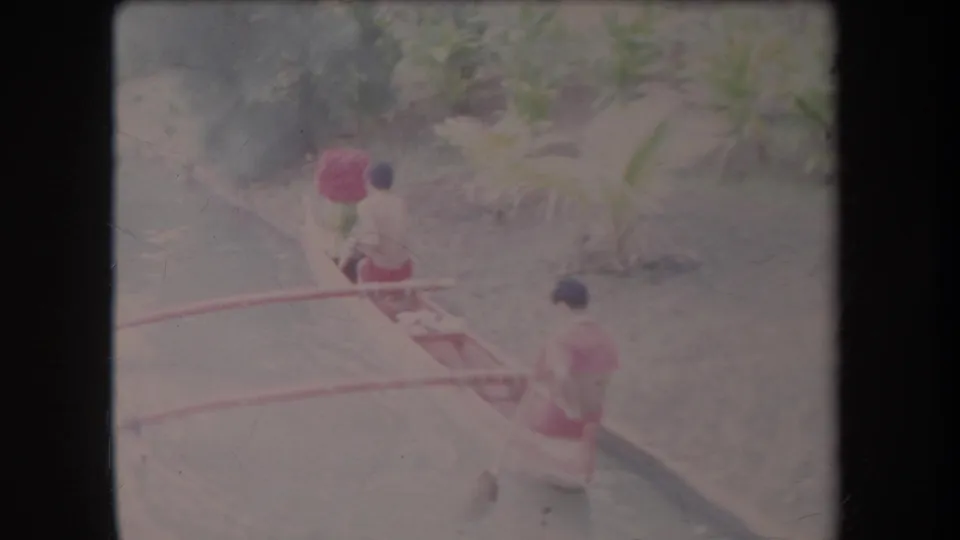 Faded 1969 Hawaii home-movie frame of two men paddling an outrigger canoe beside dense tropical plants.