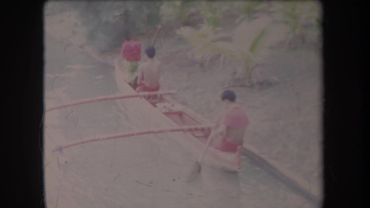 Two men paddle a small outrigger canoe along a narrow canal.