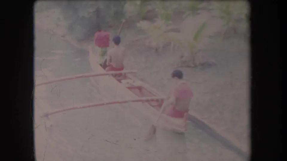 In 1969 Hawaii home-movie footage, two adult men move a small outrigger canoe through calm canal water bordered by vegetation.
