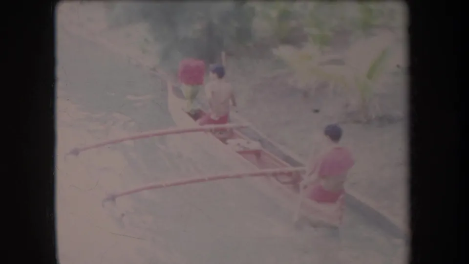 Two men in a red-accented outrigger canoe continue along a narrow canal in Hawaii, seen in soft 8mm color.