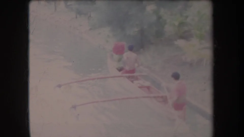 The men continue paddling the small outrigger canoe through calm water, with vegetation and soft focus surrounding the canal.