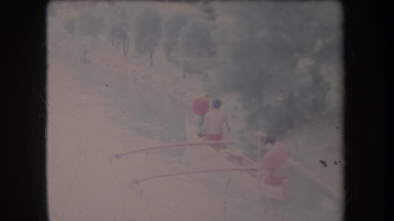 Three distant people paddle an outrigger-style canoe on pale water beside trees.