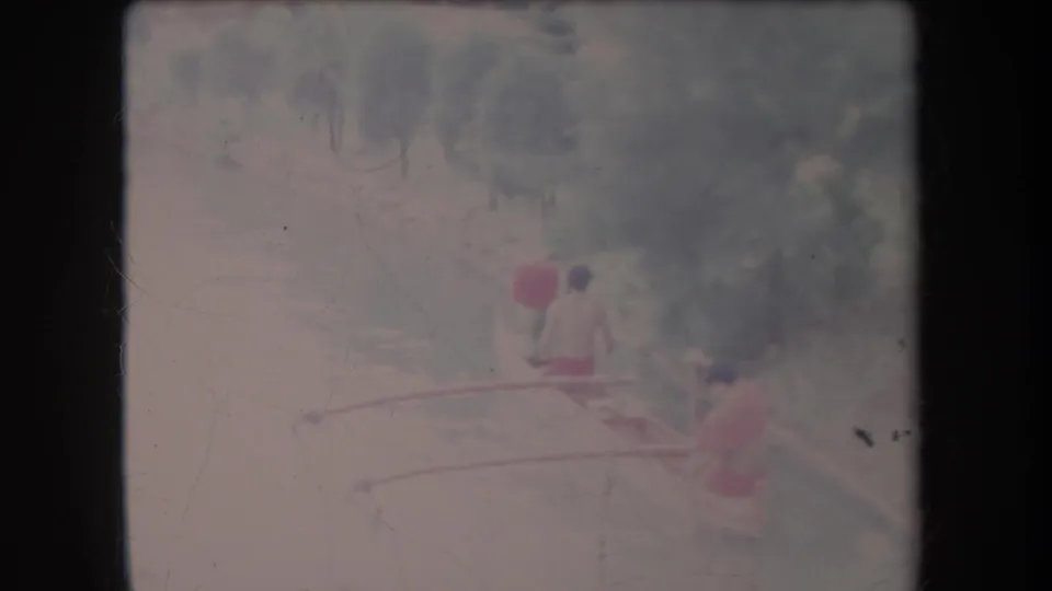 1969 Hawaii home-movie frame showing three distant paddlers in an outrigger-style canoe on a calm, tree-lined waterway.