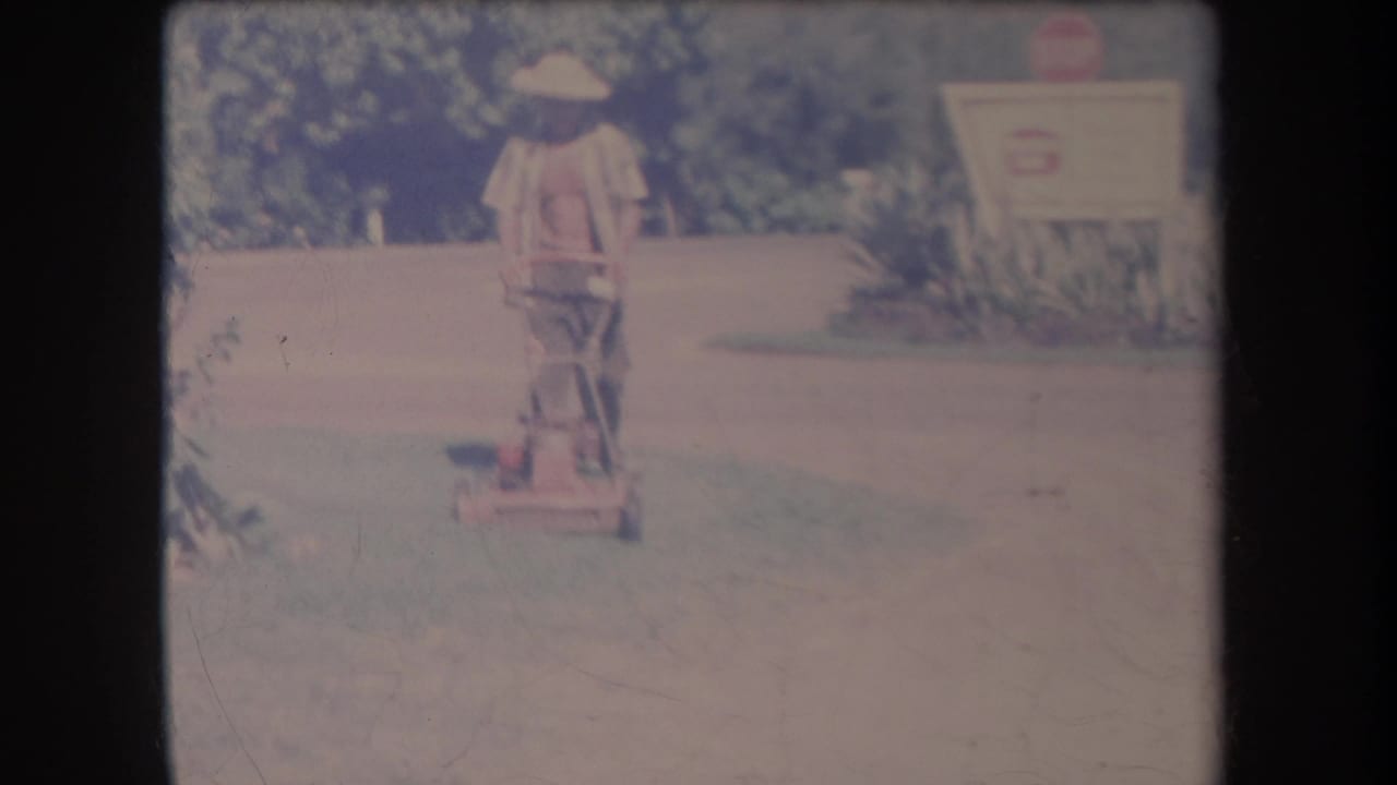 1969 Hawaii home-movie frame with a man in light clothing pushing a red mower across a grassy yard.