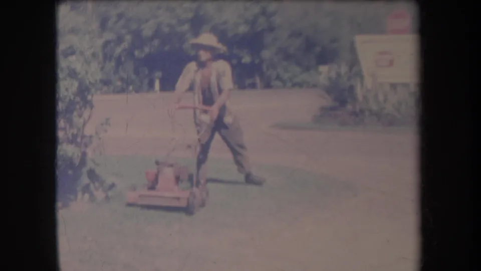 1969 Hawaii home-movie frame of the man angling the mower across the lawn beside shrubs and roadside signage.