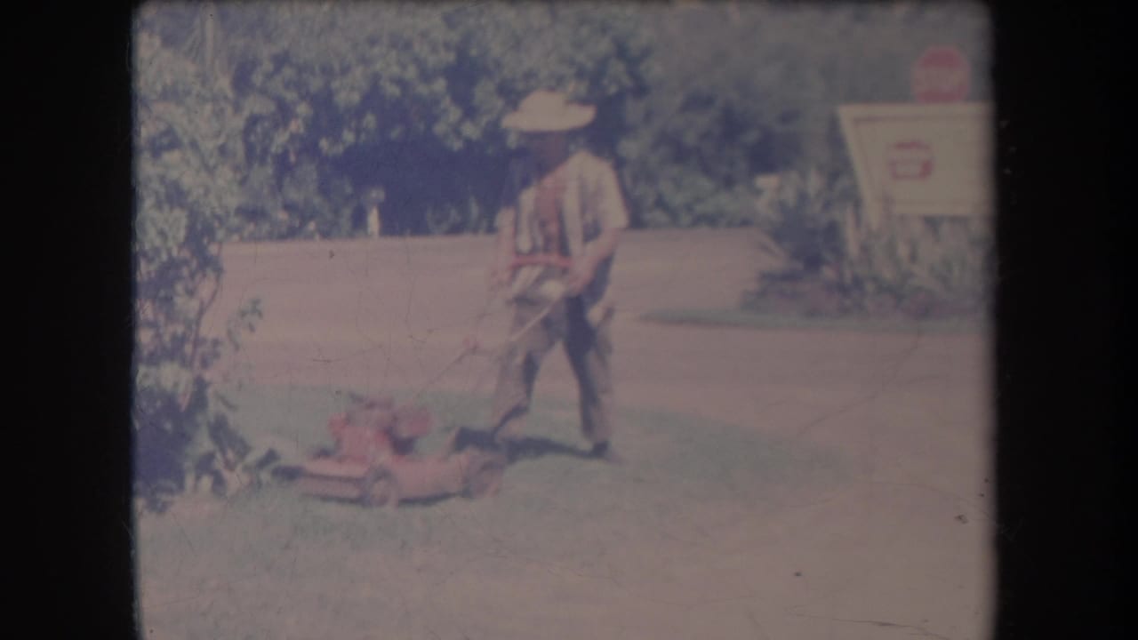 1969 Hawaii home-movie frame showing a man in a straw hat pushing a red lawn mower across grass near greenery and a roadside sign.