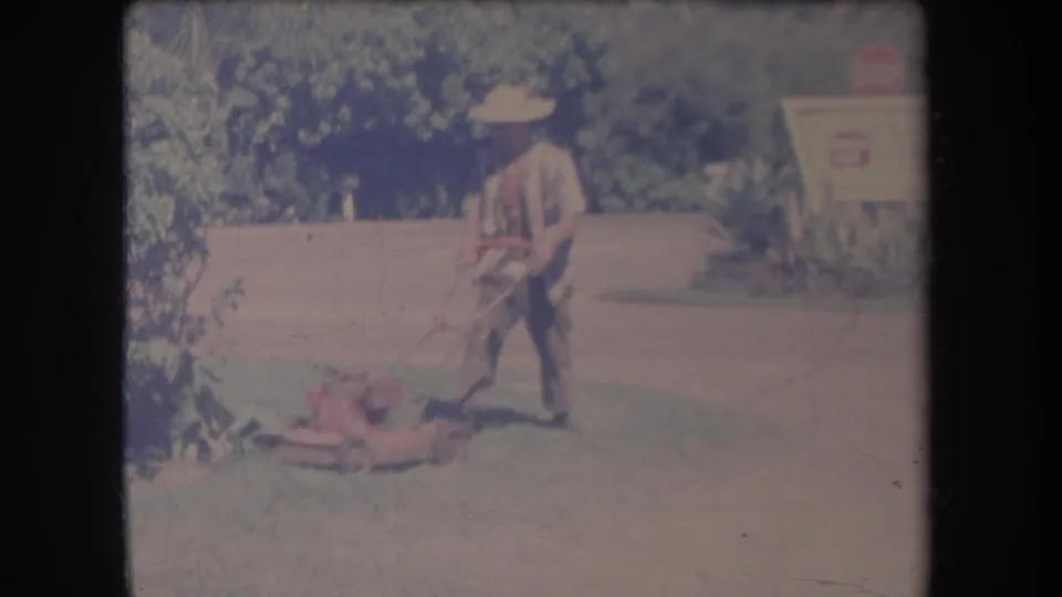1969 Hawaii home-movie frame showing a man in a straw hat pushing a red lawn mower across grass near greenery and a roadside sign.