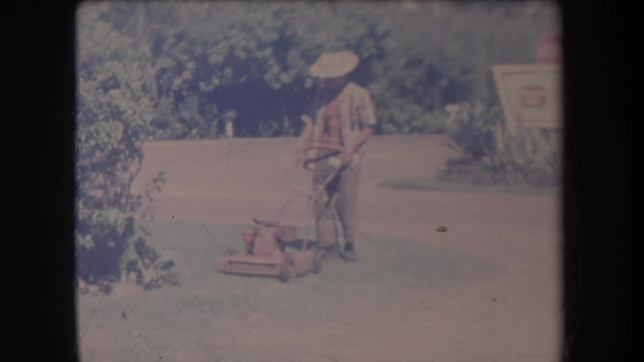 Man stands with a red push mower on a lawn beside shrubs and a sign.
