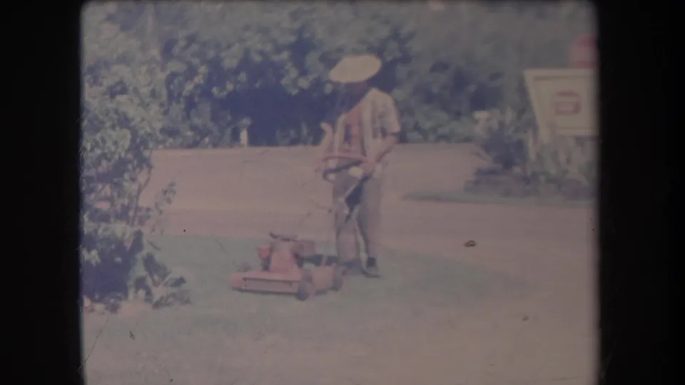 1969 Hawaii frame with a man in light clothing holding a red push mower on a lawn, with dense greenery and a sign in the background.