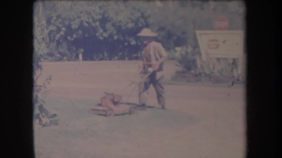 1969 Hawaii home-movie frame showing a man guiding a red push mower across the lawn, with foliage and a roadside sign beyond.