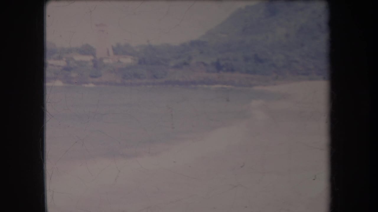 1969 Hawaii home-movie frame with a resort-like shoreline across the water, backed by trees and a hazy hillside.