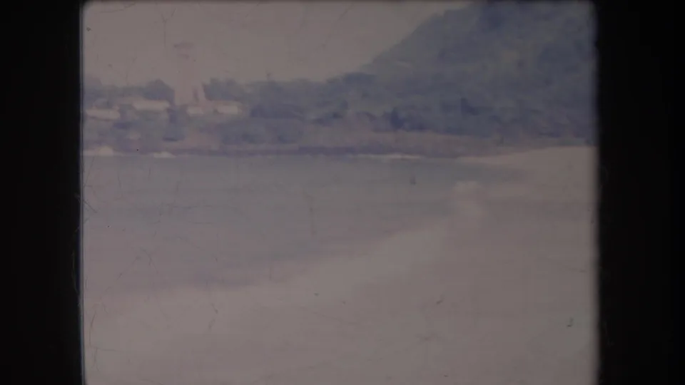 1969 Hawaii home-movie frame with a resort-like shoreline across the water, backed by trees and a hazy hillside.