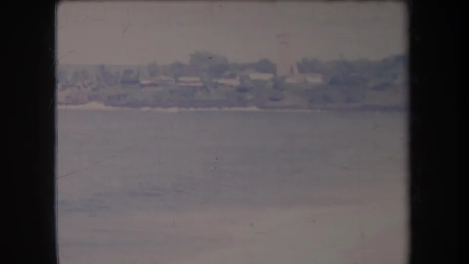 Faded 1969 Hawaii 8mm frame showing low waterfront buildings, trees, pale beach, and calm ocean water.