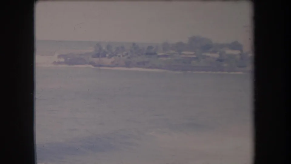 1969 Hawaii 8mm frame showing a resort shoreline, low buildings, trees, surf line, and open water in soft faded color.