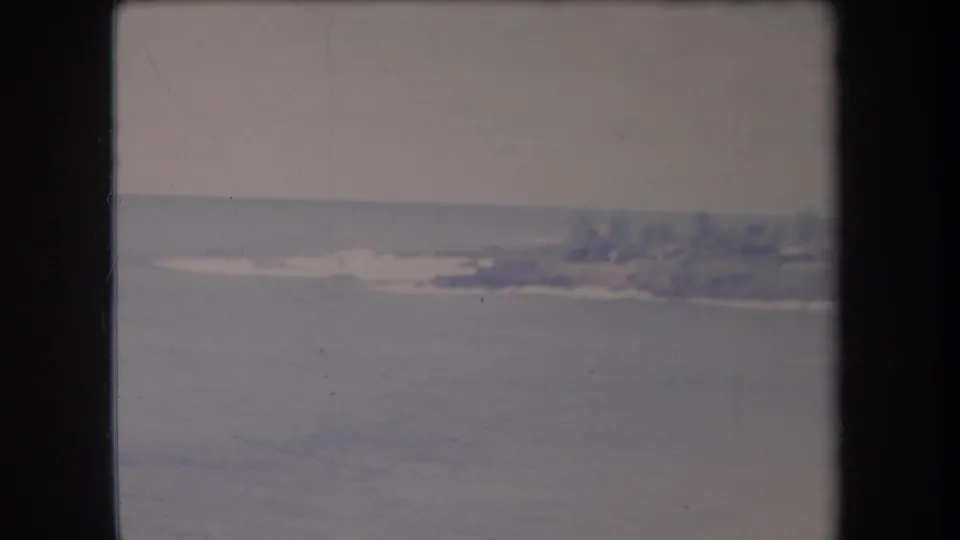 1969 Hawaii coastal home-movie frame showing open water, a hazy treed shoreline, and small low structures in the distance.