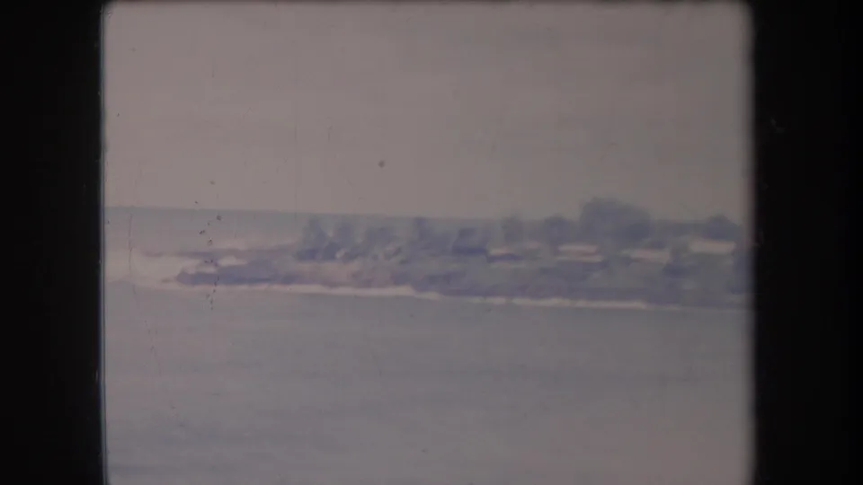 1969 Hawaii 8mm frame of a distant shoreline across blue-gray water, with trees and low buildings softened by film grain and fading.