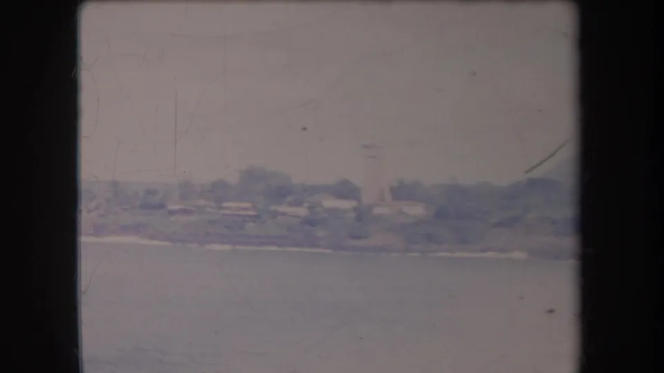 1969 Hawaii home-movie still showing calm water in the foreground and a distant coastal settlement softened by faded 8mm color.