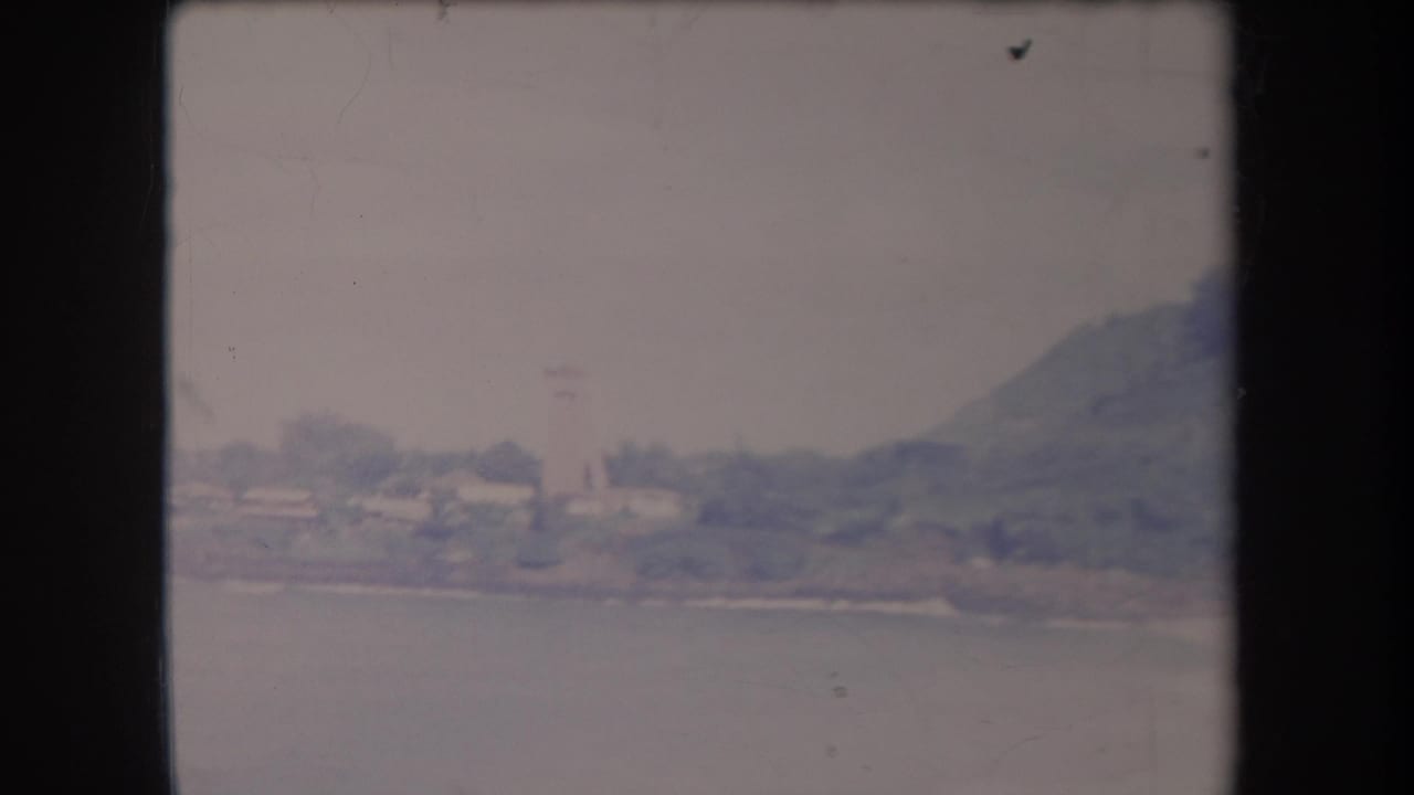 1969 Hawaii 8mm view of a distant coastal settlement, water, and a hill under a pale sky.