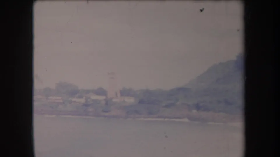1969 Hawaii 8mm view of a distant coastal settlement, water, and a hill under a pale sky.
