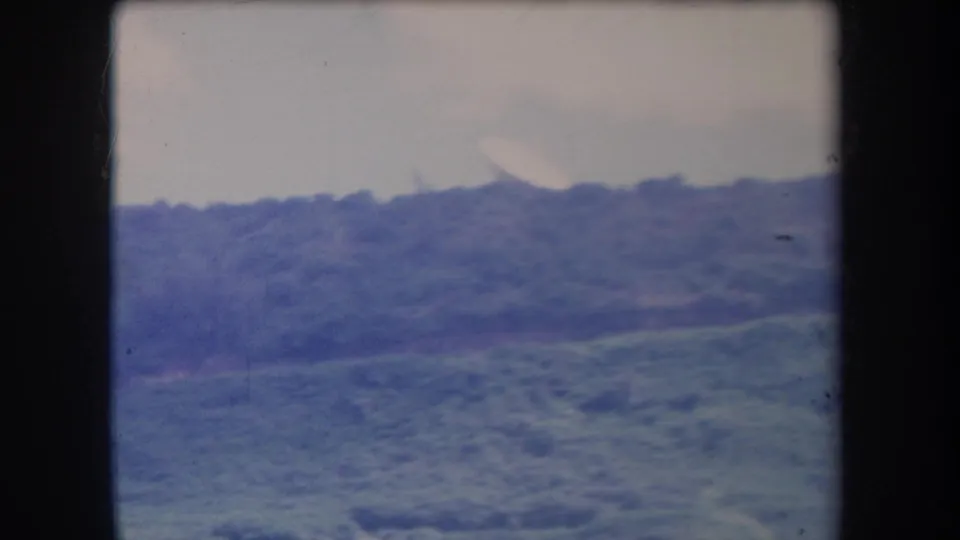 1969 Hawaii home-movie frame with a distant green slope, valley vegetation, pale sky, and visible 8mm gate edges.