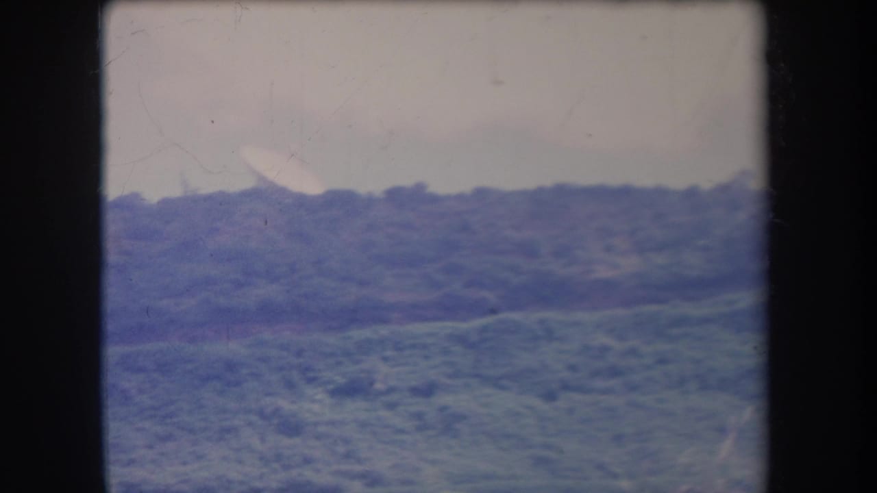 1969 Hawaii 8mm home-movie frame showing a distant foliage-covered hillside under broad daylight with soft blue-purple color shift.