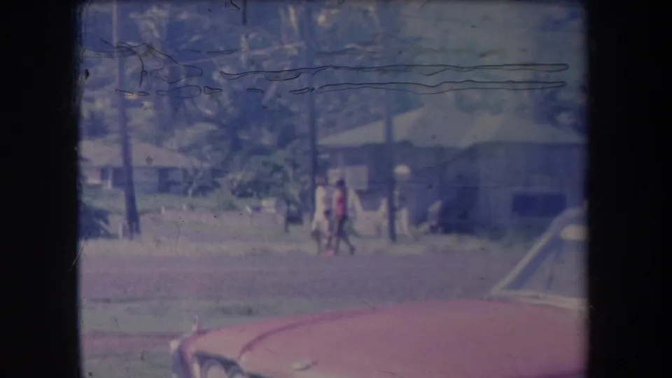 Two pedestrians walk through a Hawaii village street in 1969, with modest buildings, utility poles, palms, and a red car foreground.