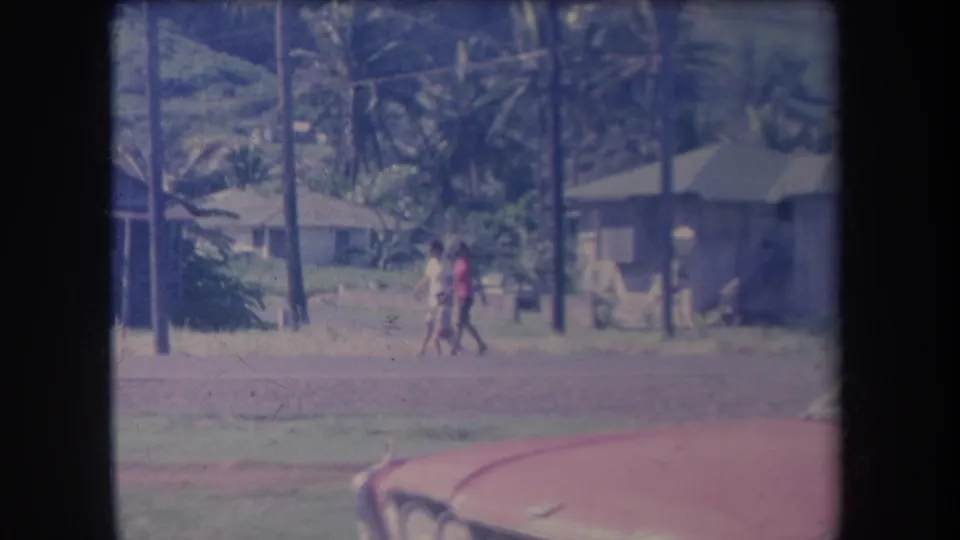 Village street view in 1969 Hawaii with two visible pedestrians, palm trees, utility poles, low buildings, and a red car in the foreground.