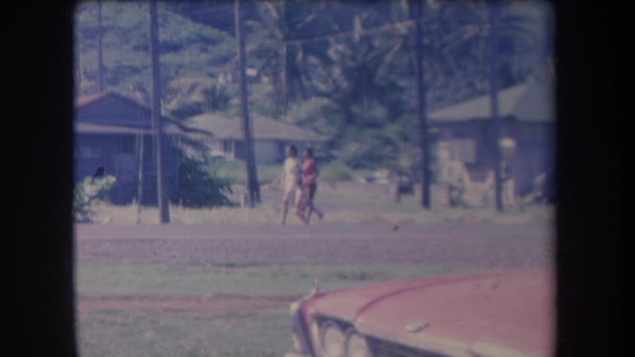 In 1969 Hawaii home-movie footage, two people walk past low houses and palm trees with a red car partly visible in front.