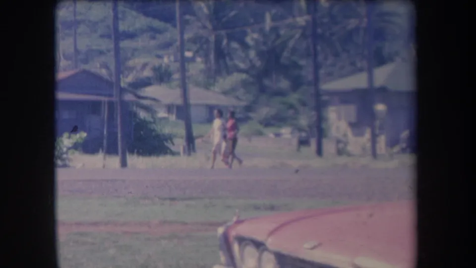 In 1969 Hawaii home-movie footage, two people walk past low houses and palm trees with a red car partly visible in front.