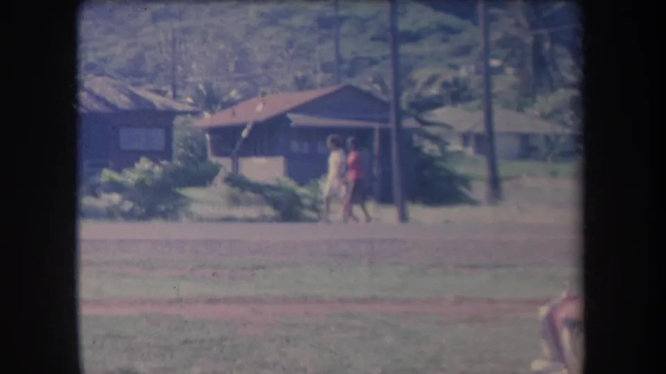 In a soft 8mm frame, two people walk past tropical shrubs and low houses in a Hawaii residential setting.
