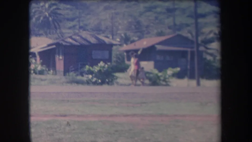 Two people walk along a quiet Hawaii roadside, framed by low houses, palms, and faded 8mm color.