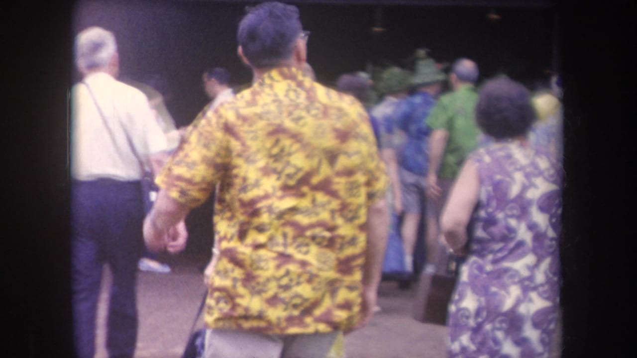 1969 Hawaii home-movie frame showing adults in bright floral clothing walking through a crowded outdoor area.