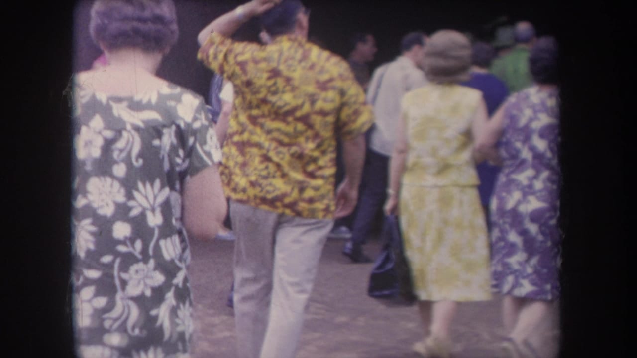 Adults in patterned dresses and a yellow floral shirt walk through a crowded walkway.