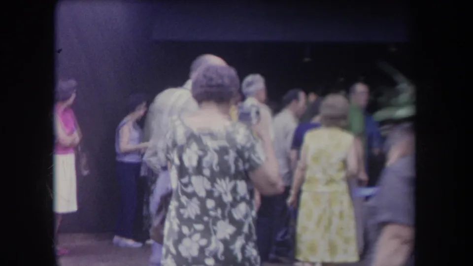 1969 Hawaii home-movie frame with adults in floral dresses moving through a crowded shaded walkway.
