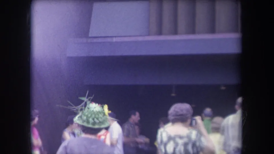 1969 Hawaii frame showing a decorative green hat in the foreground with pedestrians gathered near a shaded building.