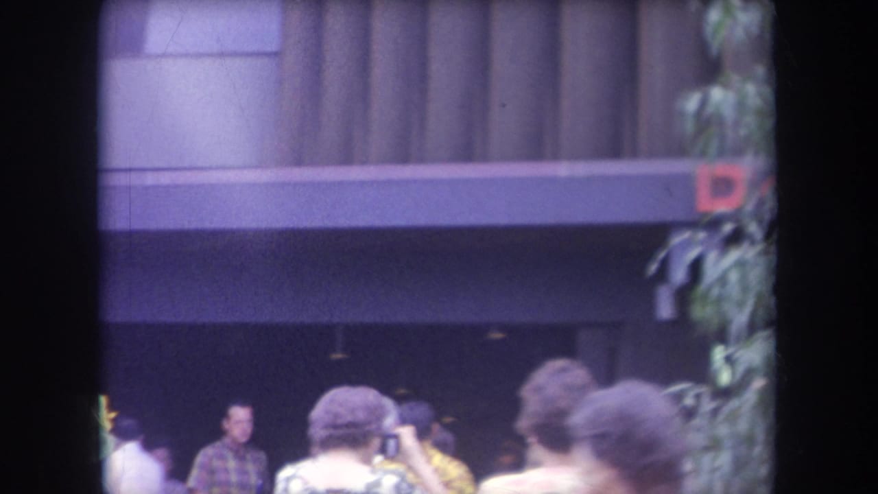 1969 Hawaii home-movie frame showing people outside a paneled building with foliage and a partial red sign letter.