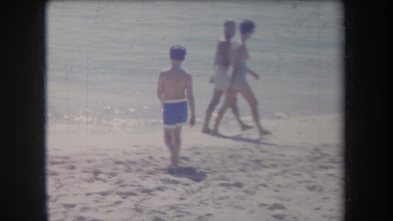1969 Hawaii beach scene with a boy in blue swim trunks facing the water and two people walking along the surf line.