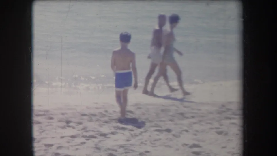1969 Hawaii beach scene with a boy in blue swim trunks facing the water and two people walking along the surf line.