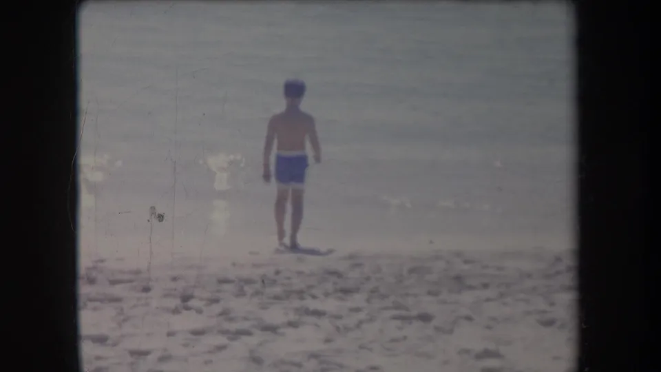 1969 Hawaii home-movie frame of a boy in blue swim trunks on sand, seen from behind near calm ocean water.