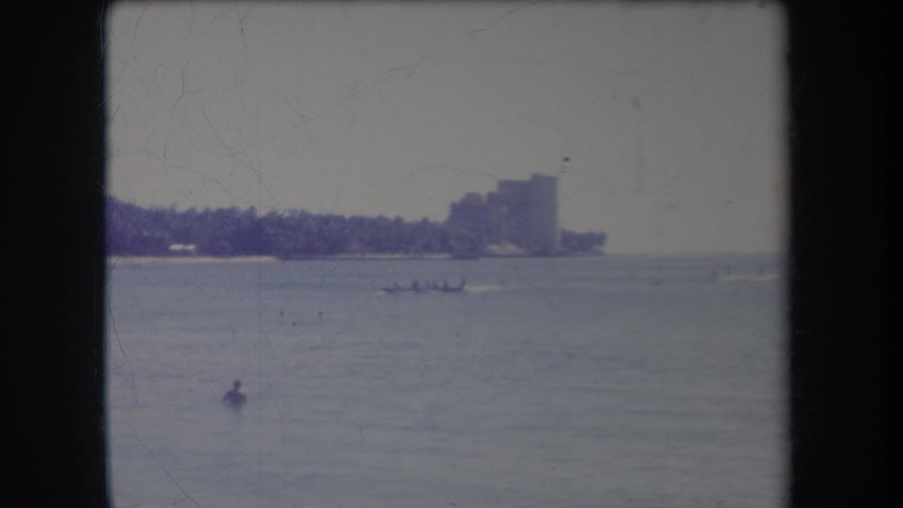 1969 Hawaii 8mm frame showing calm coastal water, a distant palm-lined shore, a large building, small craft, and a swimmer.